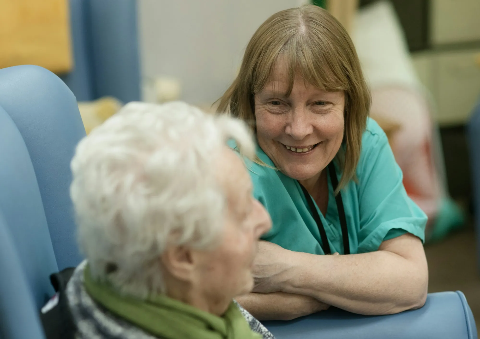 A care worker in green scrubs leaning in to talk with an older person at home