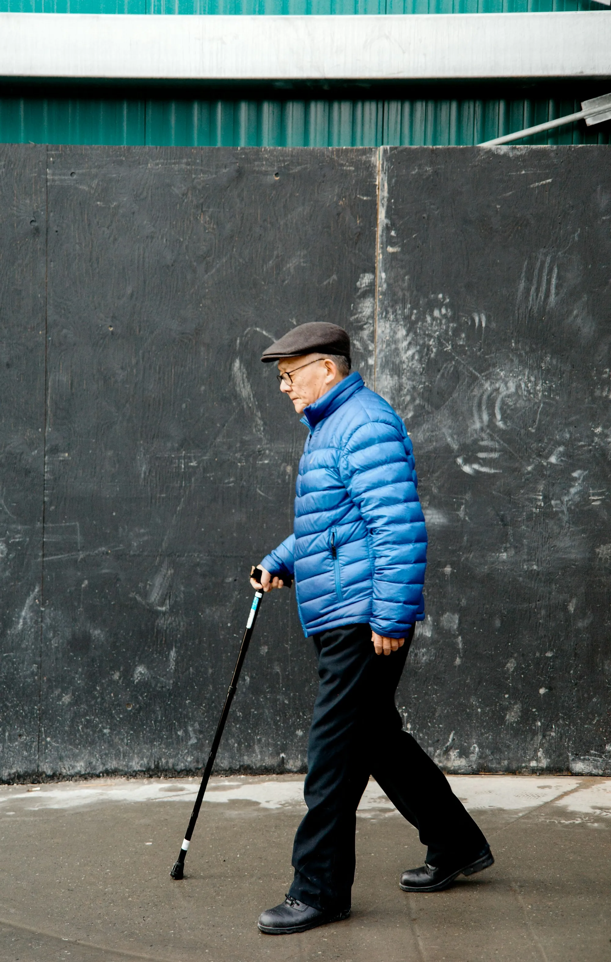 An older man walking a hallway with a wheeled frame while an aide walks beside him